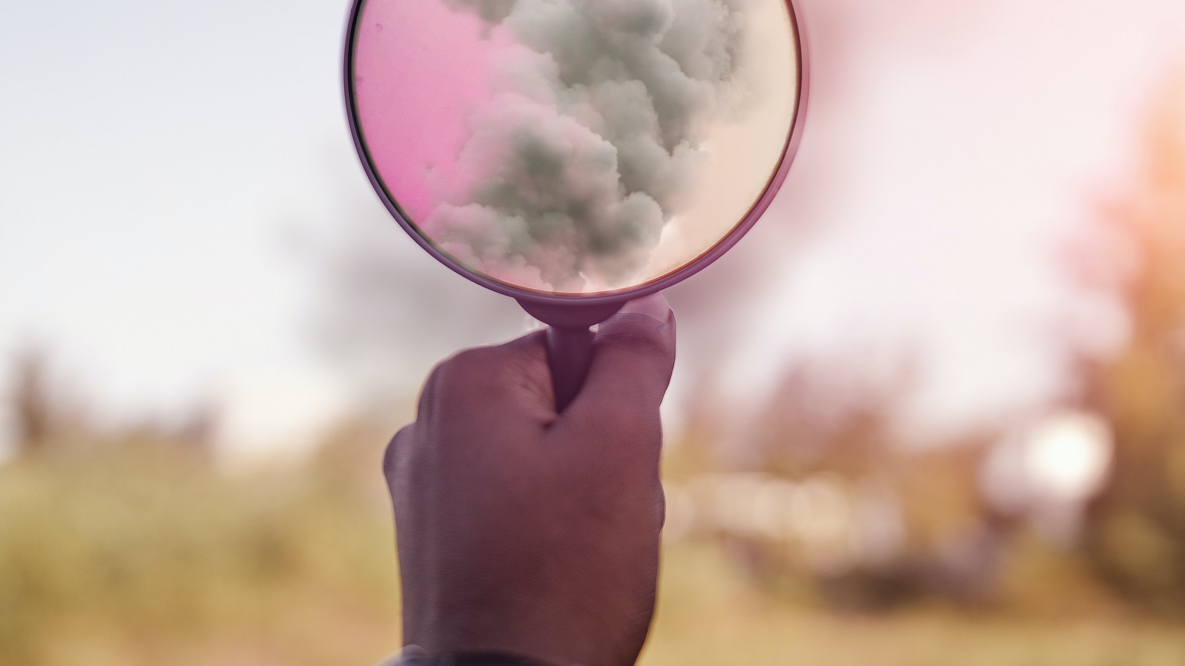 Cover Photo: This picture shows someone holding up to a magnifying glass filled with smoke. The smoke becomes pink and magical looking through the magnifying glass.