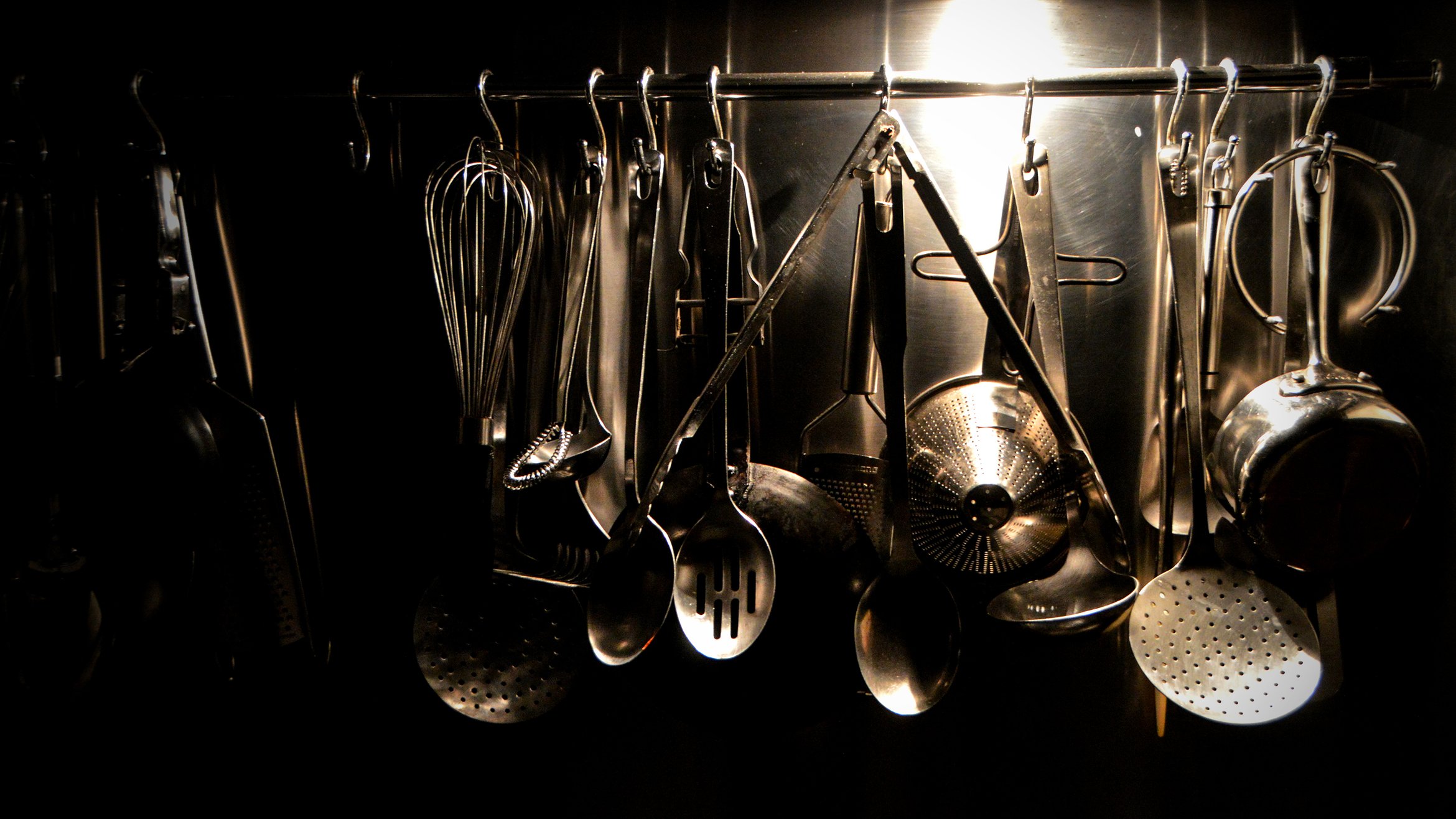 Cover Photo: culinary tools like whisks, strainers, pots, and pans hanging in a dark kitchen, lit by an ominous light