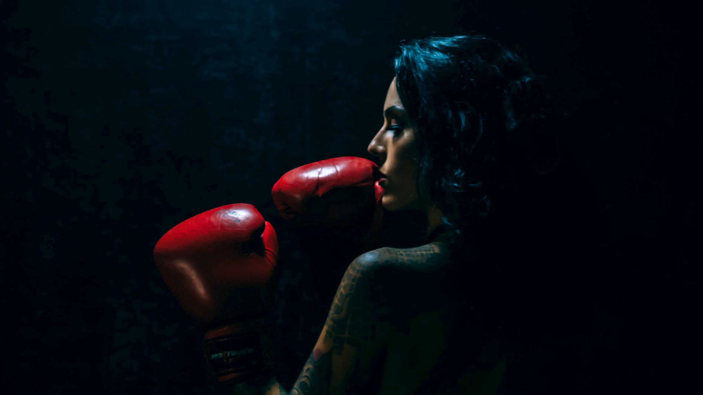 Cover Photo: A photograph of a femme person wearing a red pair of boxing gloves and raising it towards their face. They appear in profile against a dark background.