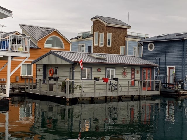 This photograph shows a cute gray house floating in a wharf. It has window boxes and a bike sitting on its porch. Other various floating homes in oranges, blues, and browns, are behind it.