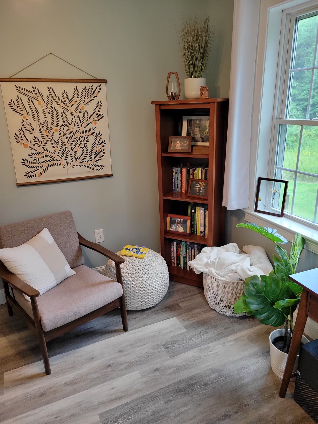 In this photograph, we see a slim book shelf, a wall hanging, and an armchair propped so that it can see out of a window